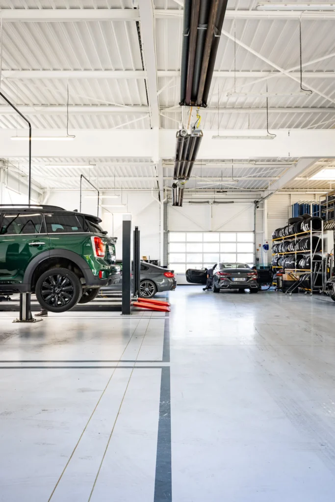 Intérieur d'un atelier automobile avec plusieurs voitures, un véhicule vert sur un pont élévateur et des étagères de pneus sur le côté droit.
