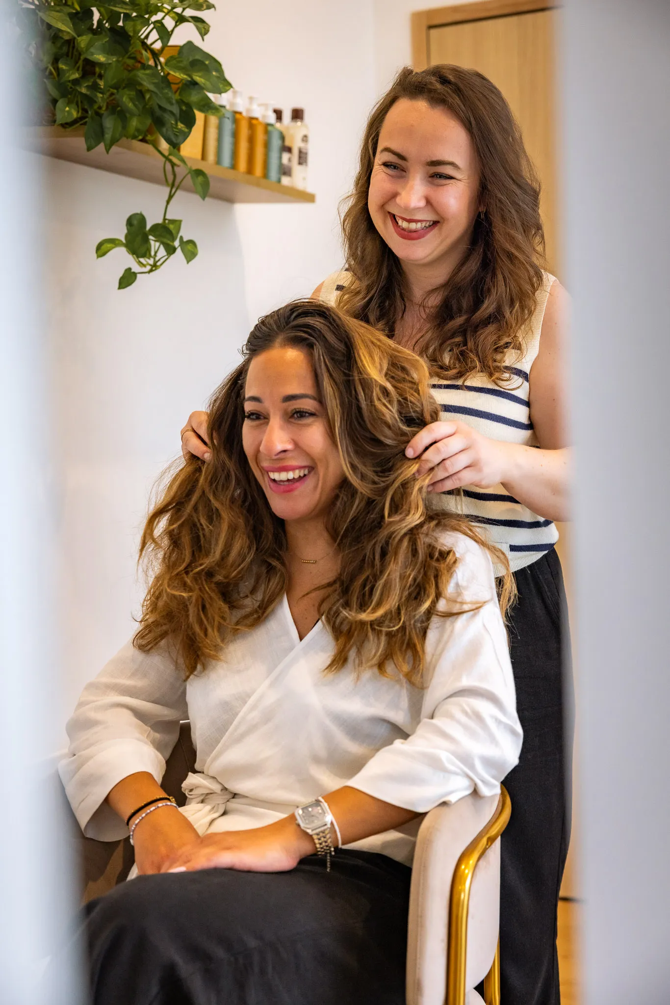 Une coiffeuse souriante coiffant une femme assise avec de longs cheveux bouclés dans un salon.