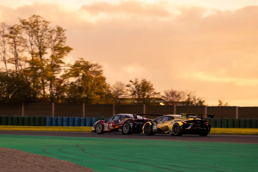 Deux voitures de course en compétition sur une piste avec un ciel orange au coucher du soleil et des arbres en arrière-plan