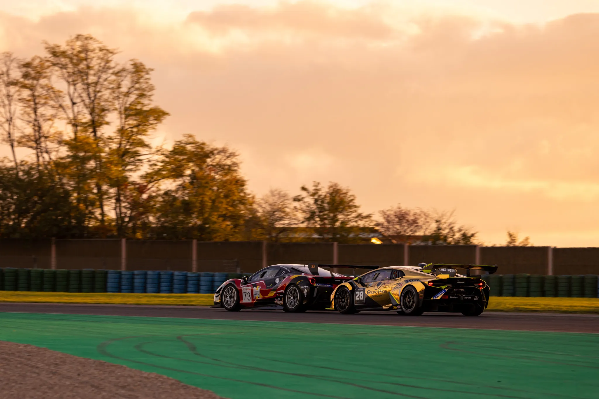 Deux voitures de course en compétition sur une piste avec un ciel orange au coucher du soleil et des arbres en arrière-plan
