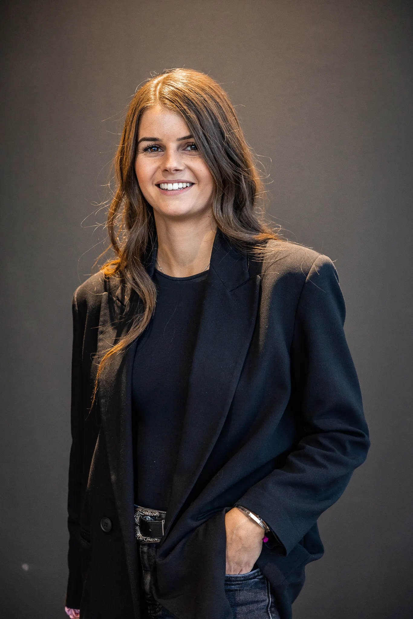 À propos Femme souriante aux cheveux longs portant une veste noire et un t-shirt noir, posant devant un fond gris.
