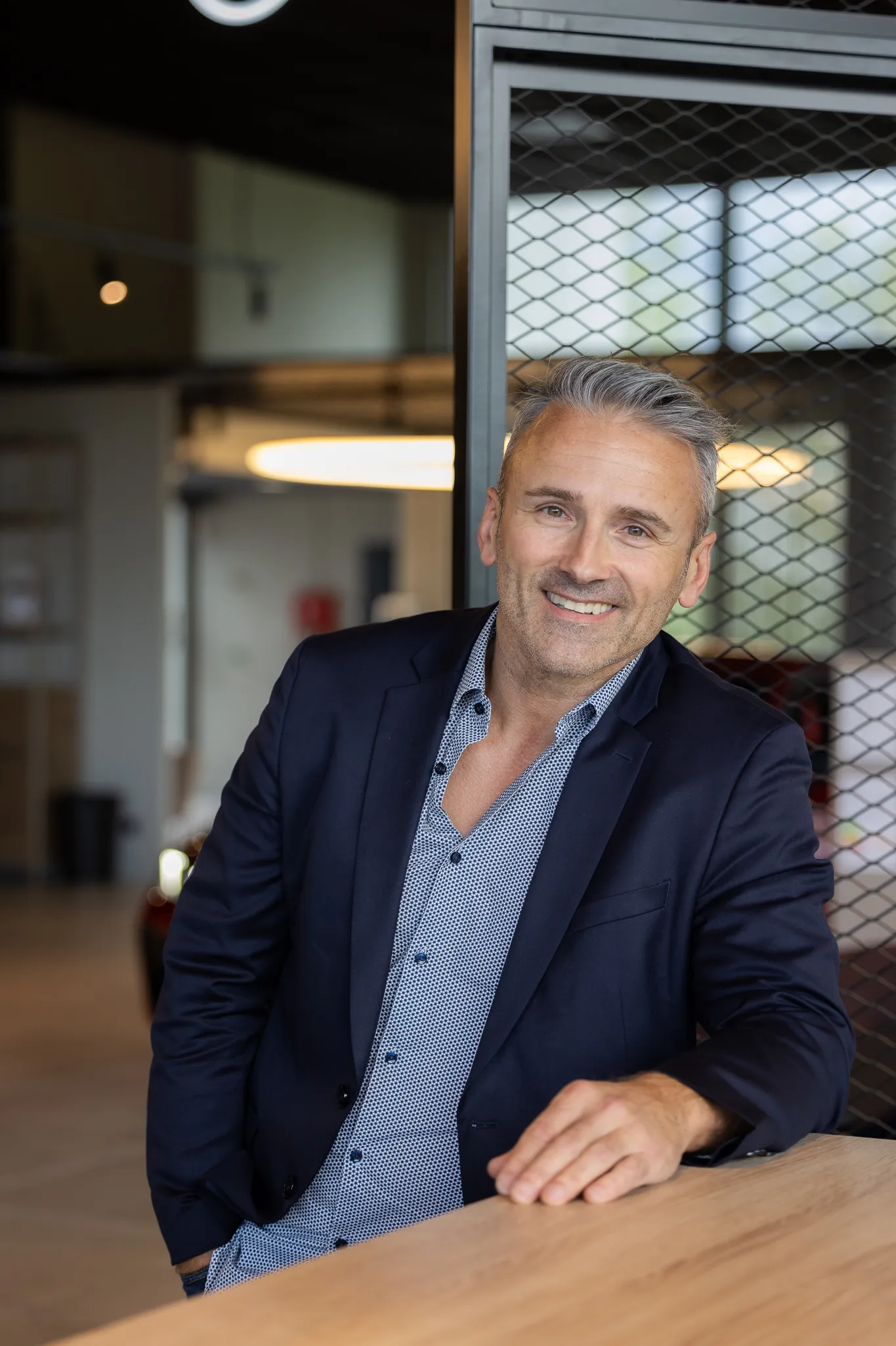 Homme souriant en chemise bleue à motifs et veste noire appuyé sur un comptoir en bois dans un intérieur moderne