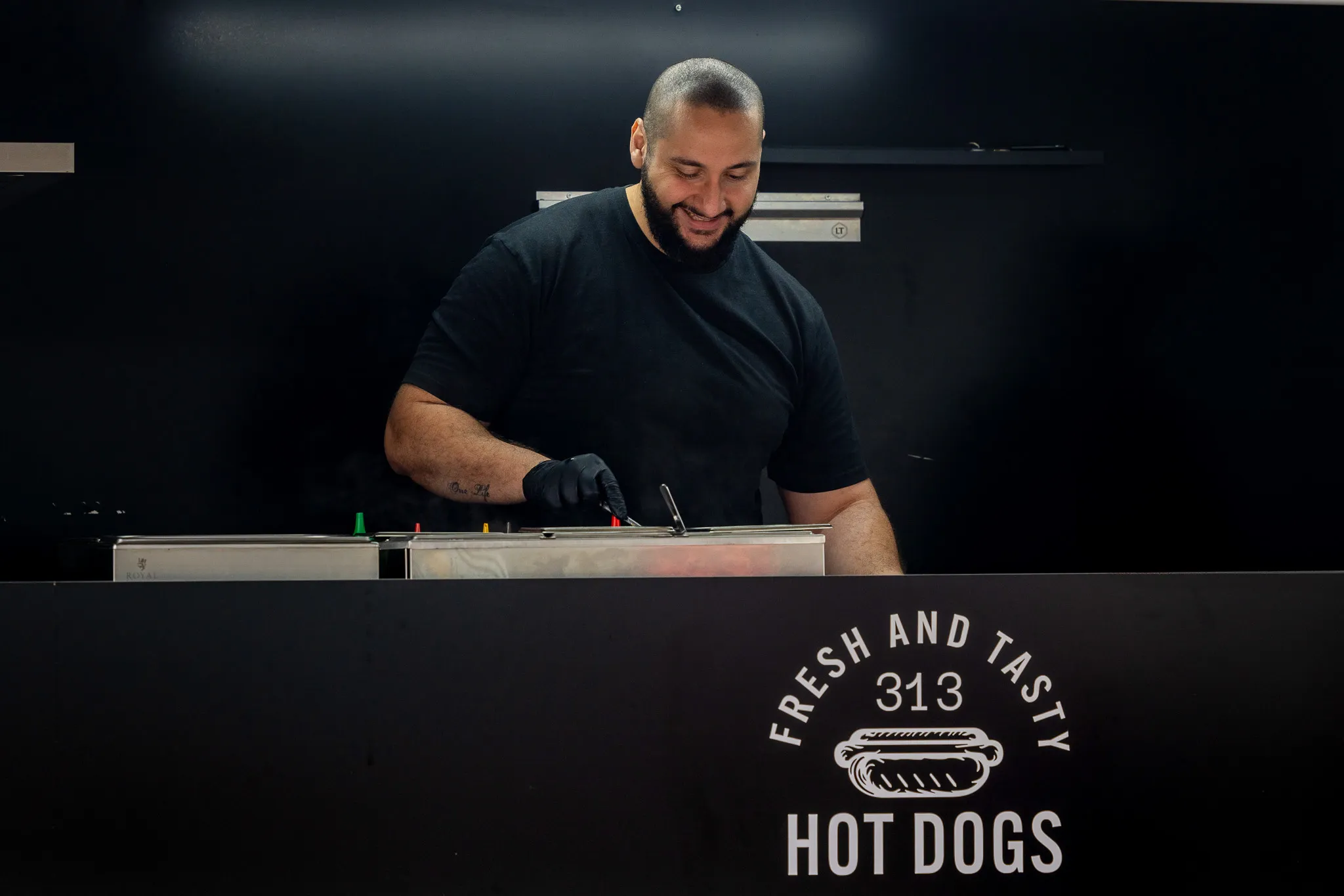 Un homme souriant portant un t-shirt noir et un gant sert des hot dogs dans un stand avec l'inscription 'Fresh and Tasty 313 Hot Dogs'.