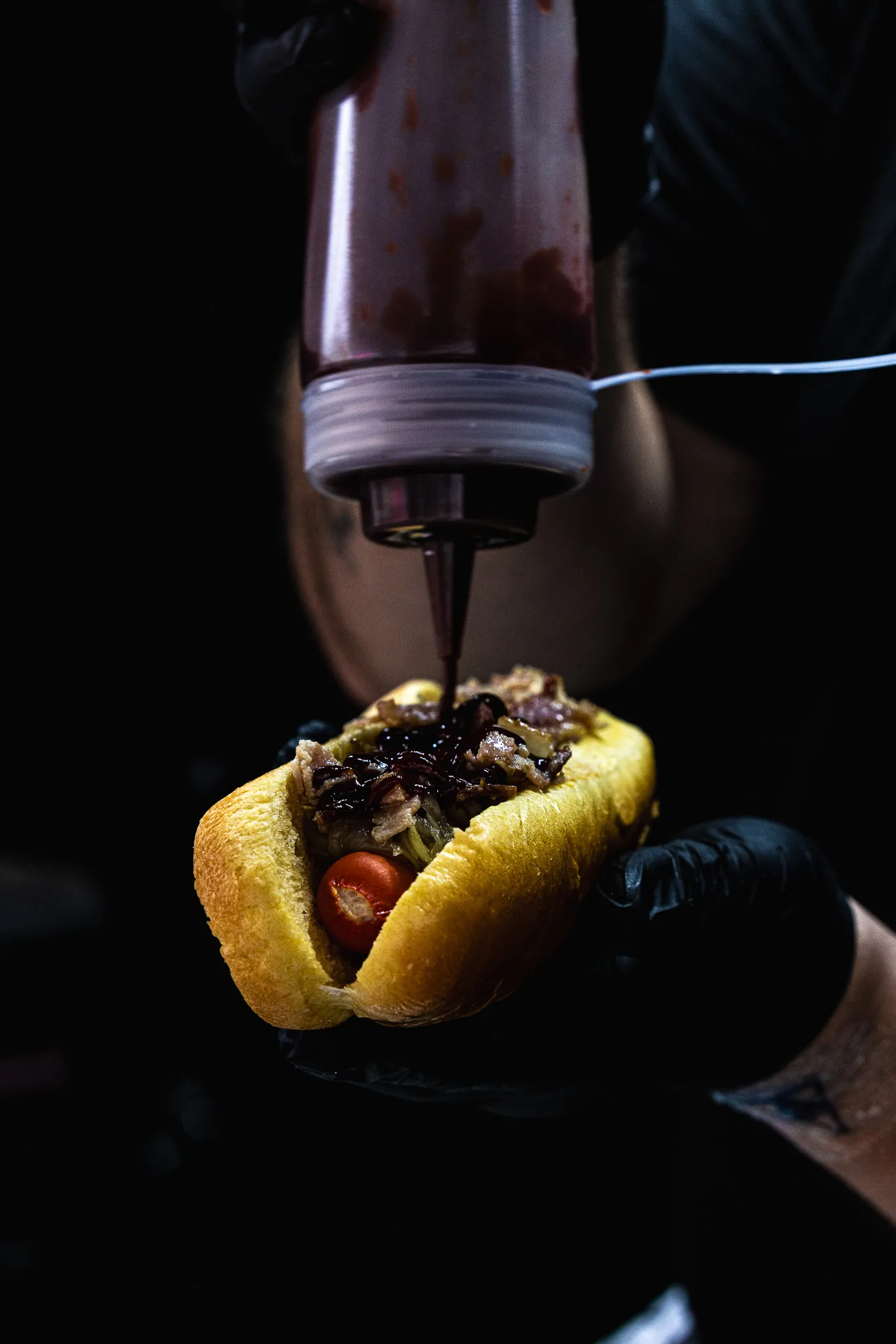 Main gantée tenant un hot dog avec une saucisse, garniture et sauce noire versée depuis une bouteille