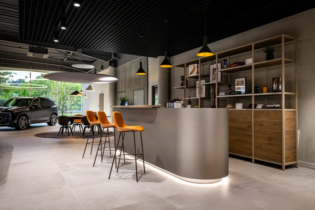 Intérieur d'un showroom avec un bar gris, quatre chaises hautes orange, une étagère en bois et une voiture noire visible à l'arrière.