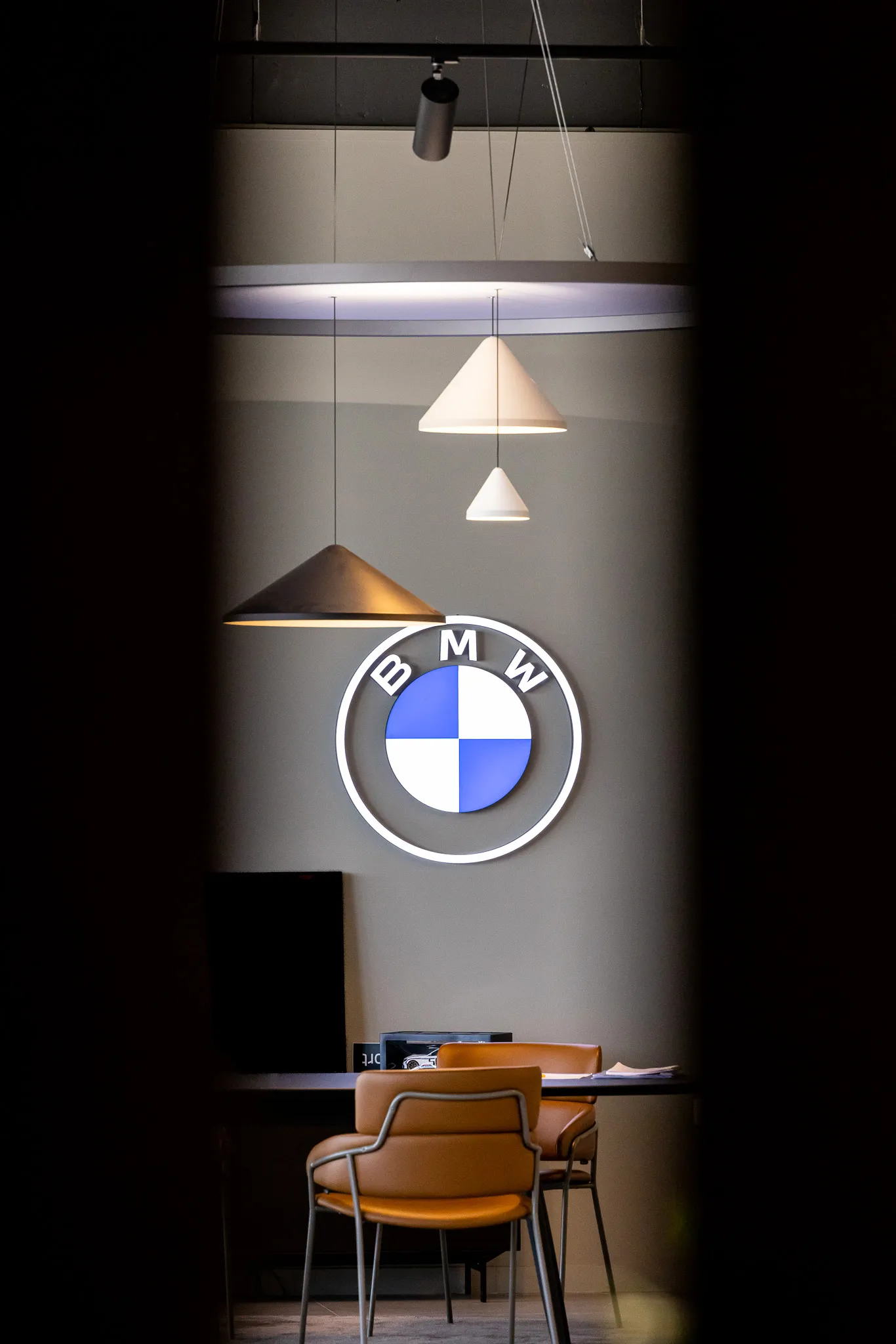 Logo BMW lumineux sur un mur gris avec trois lampes suspendues et deux chaises en cuir devant un bureau