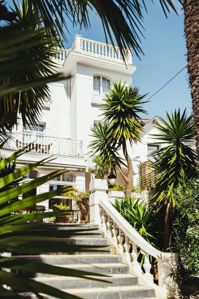 Maison blanche à plusieurs étages avec un escalier en pierre et des palmiers autour sous un ciel bleu clair
