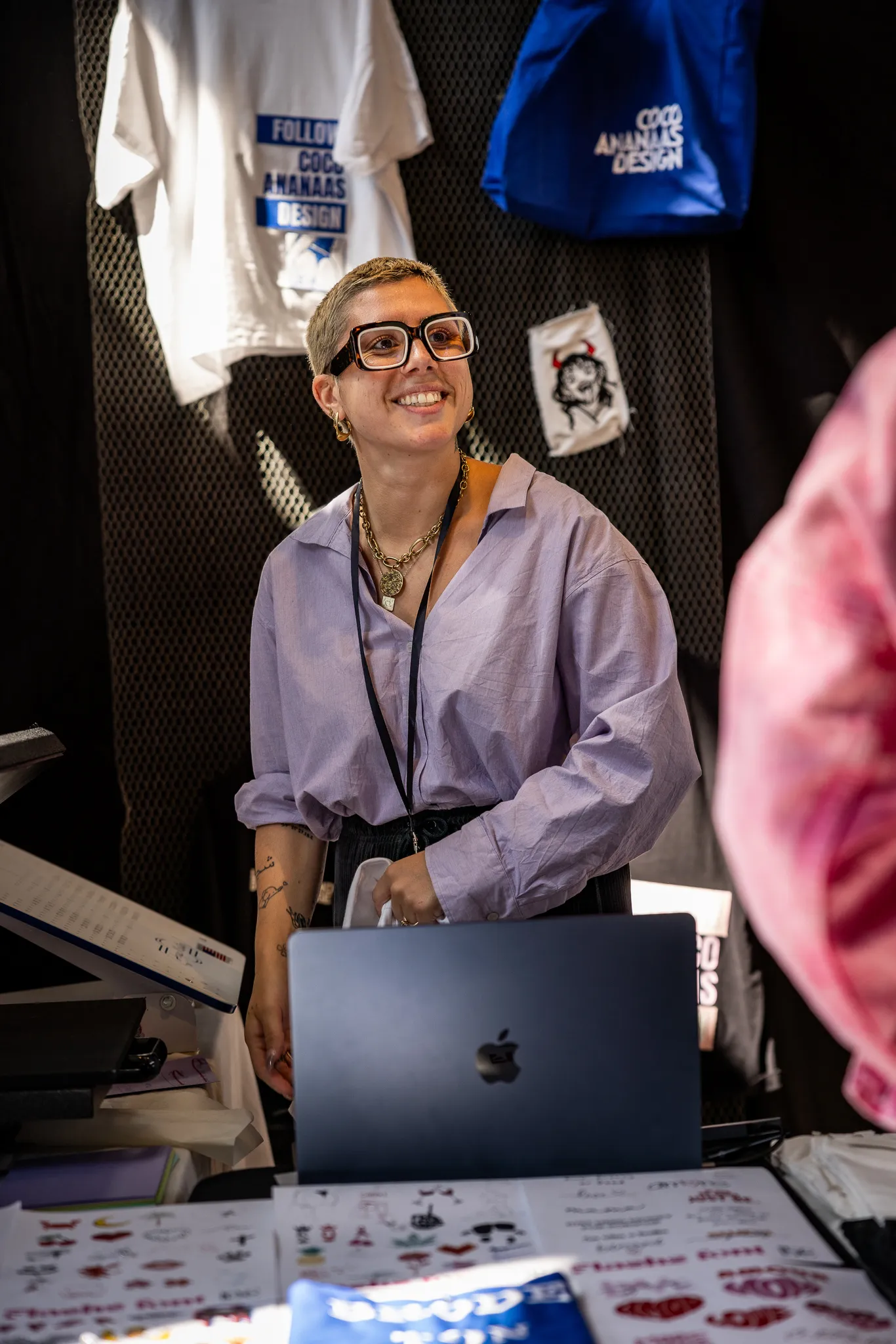 Personne souriante portant une chemise lilas et de grandes lunettes, debout devant un ordinateur portable Apple avec des vêtements suspendus en arrière-plan.