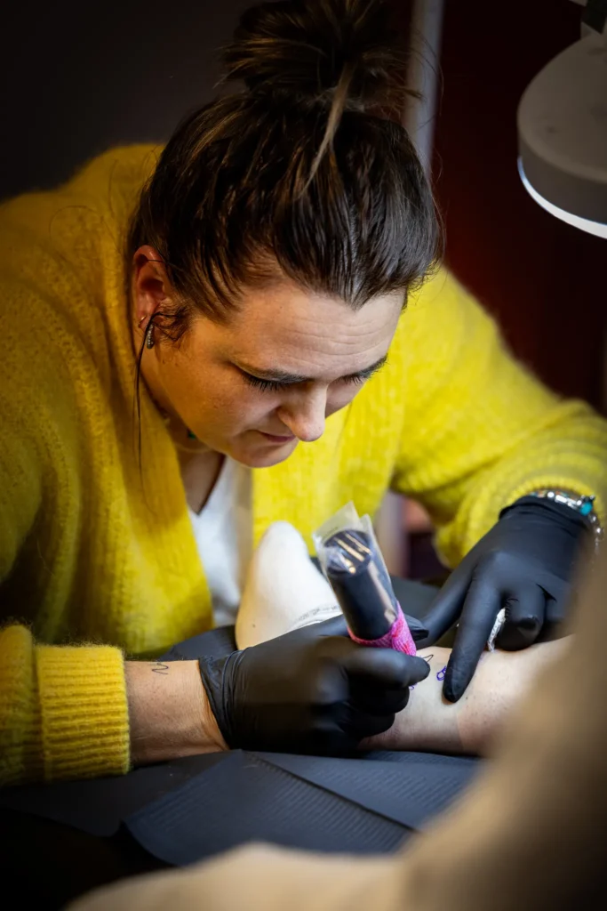 Une personne portant un pull jaune réalise un tatouage sur le bras d'un client en utilisant une machine à tatouer.