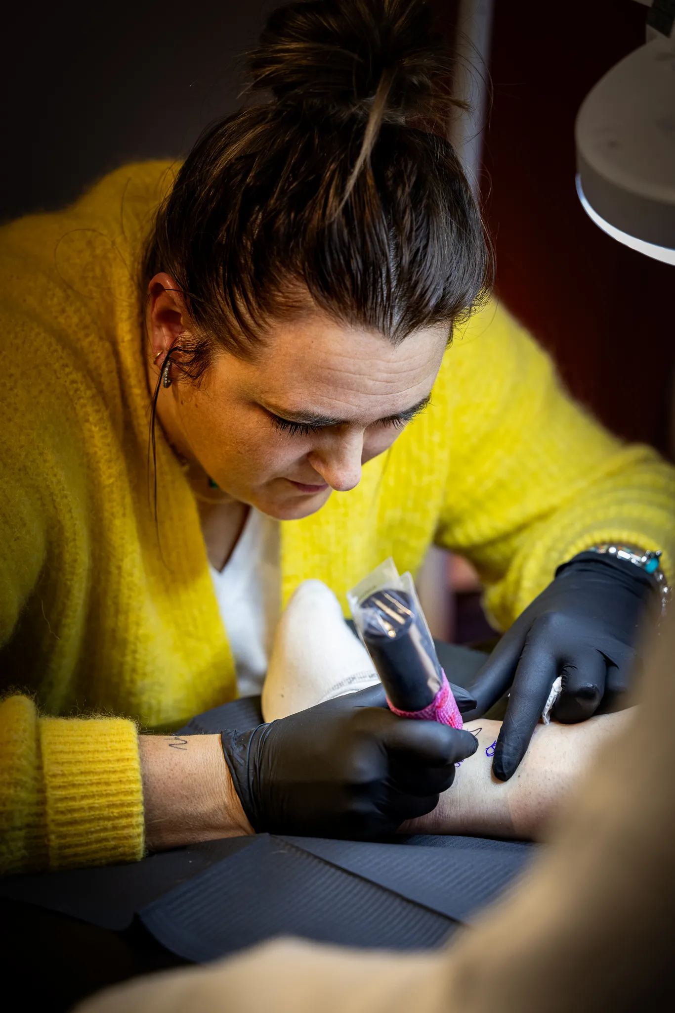 Une personne portant un pull jaune réalise un tatouage sur le bras d'un client en utilisant une machine à tatouer.