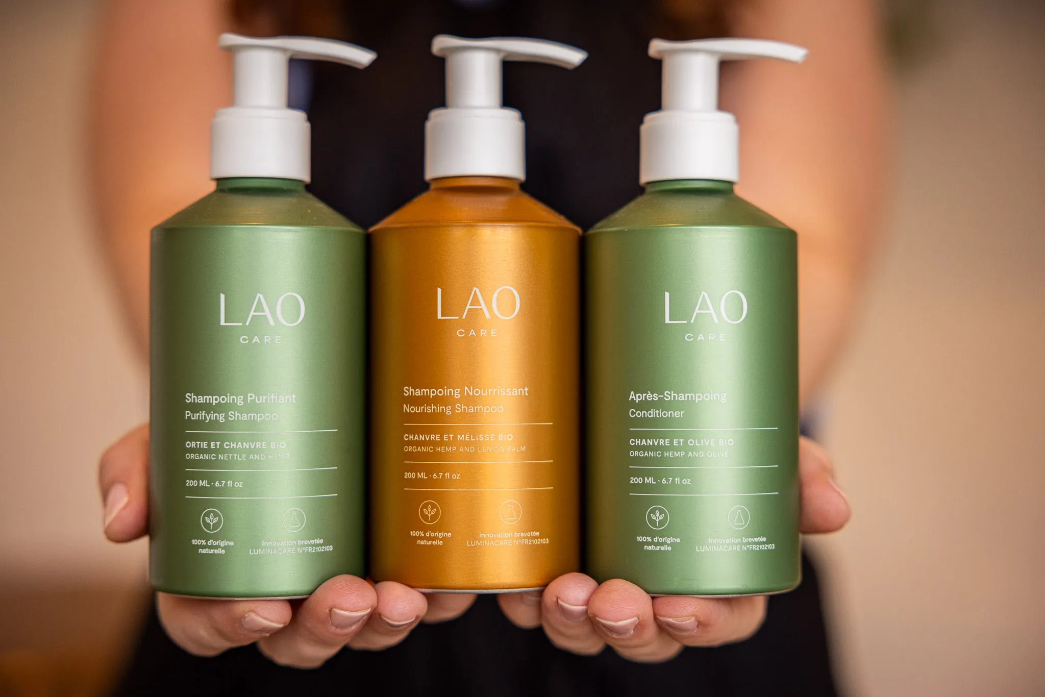 Trois flacons de soins capillaires Lao Care avec pompe, deux verts et un orange, tenus par une personne.