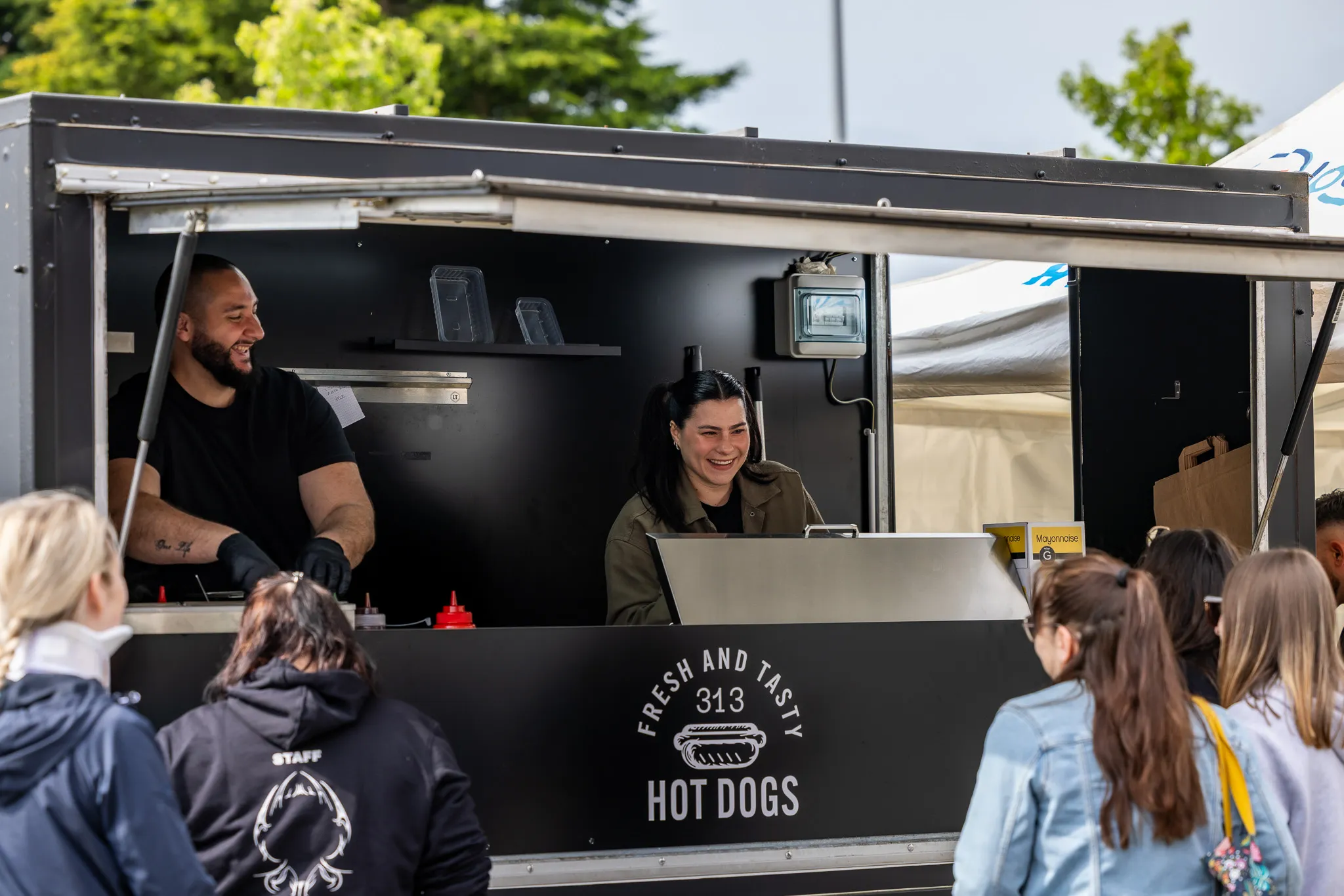 Deux vendeurs souriants dans un stand de hot dogs noir avec plusieurs clients devant eux en extérieur