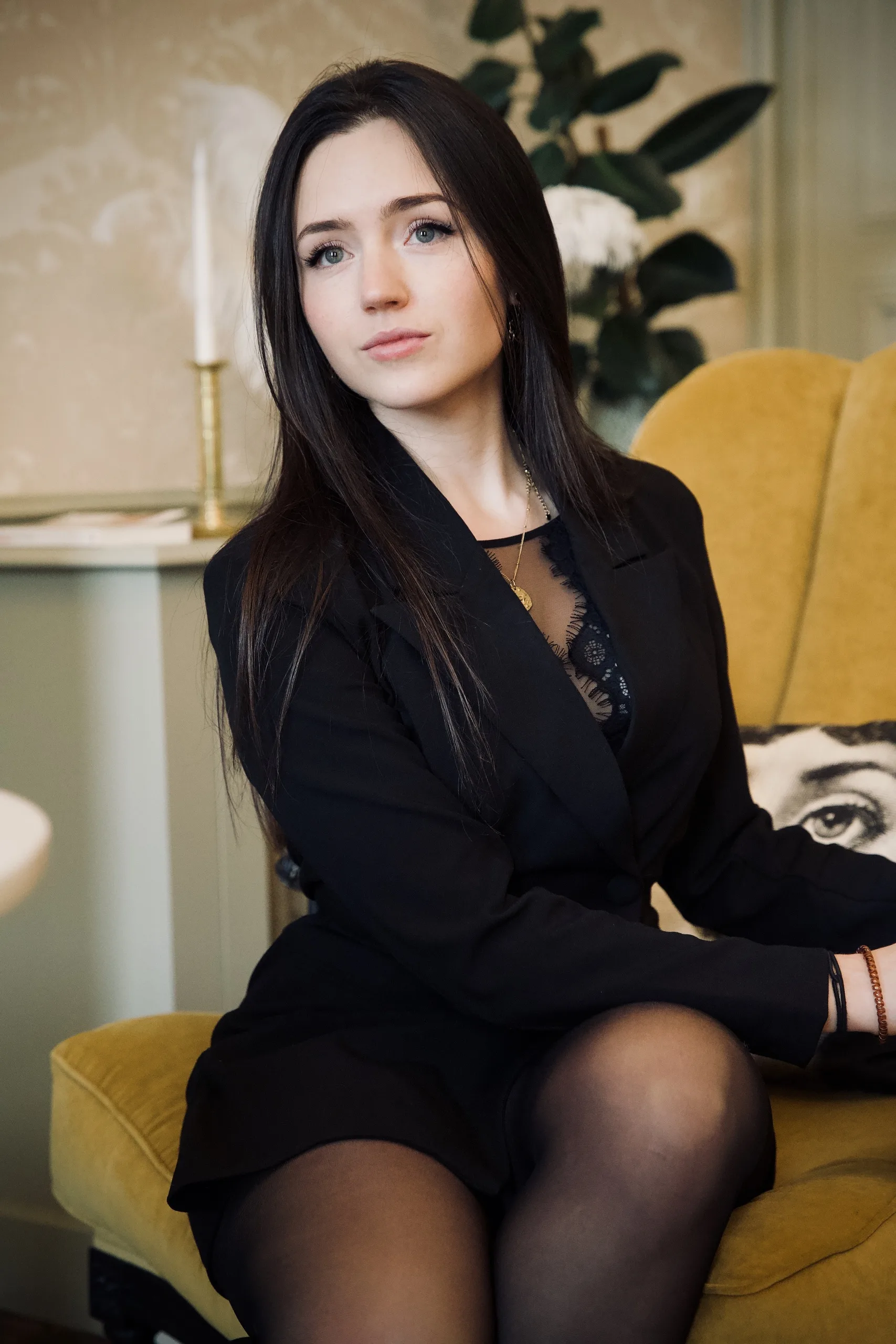 À propos Alexandra, vêtue d'un blazer noir, assise sur un fauteuil jaune dans un intérieur décoré.